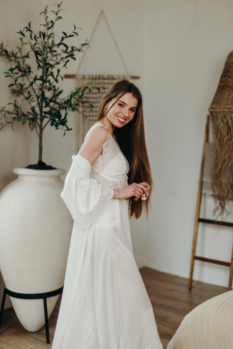 May include: A white satin bridal robe and gown with lace detailing. The gown has a V-neckline and a flowing skirt. The robe has wide sleeves. The image is taken indoors with a decorative plant and ladder in the background.