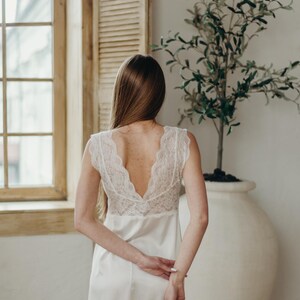 May include: A white satin nightdress featuring a lace bodice. The nightdress has a V-neck and cap sleeves. The person is standing in front of a window and a large white vase with a small tree.