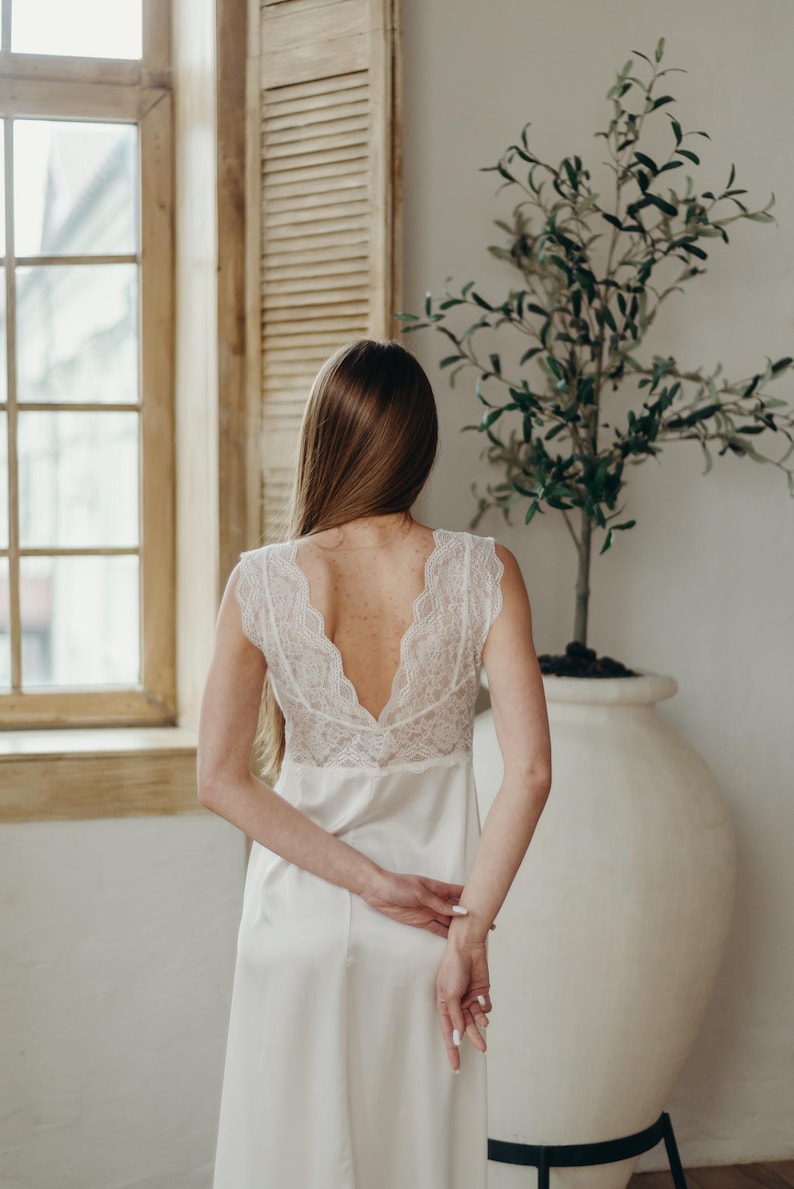 May include: A white satin nightdress featuring a lace bodice. The nightdress has a V-neck and cap sleeves. The person is standing in front of a window and a large white vase with a small tree.