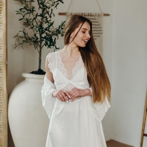 May include: A white satin lingerie set with lace detailing. The set comprises a long gown and a matching robe. The model is smiling, with long brown hair. The background includes a large white vase with a plant.