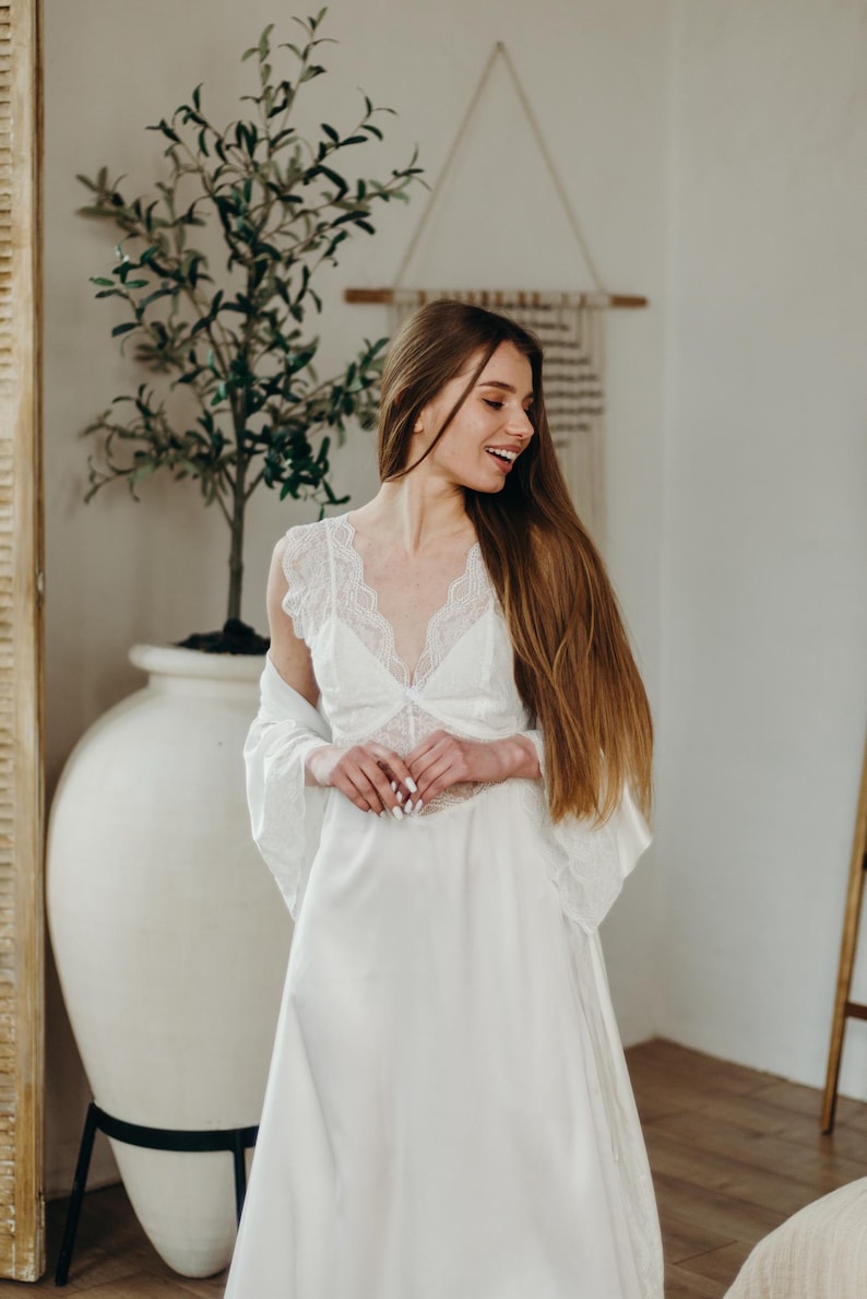 May include: A white satin lingerie set with lace detailing. The set comprises a long gown and a matching robe. The model is smiling, with long brown hair. The background includes a large white vase with a plant.