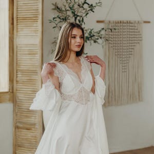 May include: A white satin bridal robe with lace detailing on the bodice and sleeves. The robe has a deep V-neckline and a flowing skirt. The sleeves are flared, and the robe is open in the front. The image is of a woman wearing the robe.
