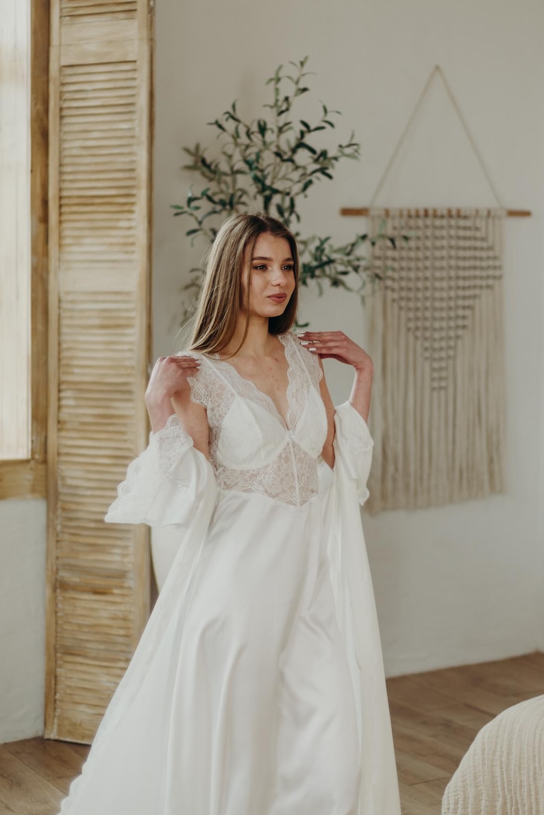 May include: A white satin bridal robe with lace detailing on the bodice and sleeves. The robe has a deep V-neckline and a flowing skirt. The sleeves are flared, and the robe is open in the front. The image is of a woman wearing the robe.