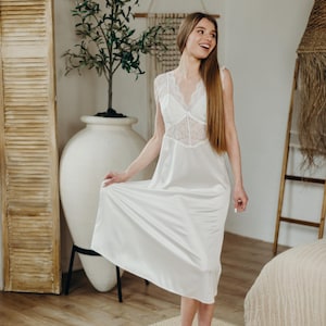 May include: A white satin slip dress with a lace bodice and a V-neck. The dress is knee-length and sleeveless. The model is wearing the dress and smiling. The background includes a large white vase with a plant and a wooden shutter.