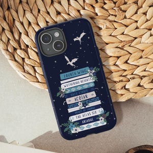 May include: A navy blue phone case with a design of stacked books, including "Fourth Wing" and "Wuthering Heights", decorated with floral and dragon motifs. The phone case rests on a woven mat, alongside a notebook and a small wooden bowl.