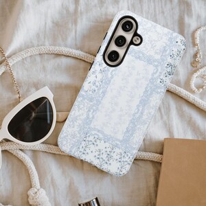 Op de afbeelding: Een rechthoekig smartphonehoesje met een lichtblauw en wit bloemmotief en een centraal rechthoekig kader. Het hoesje heeft een zwarte uitsparing voor de cameramodule met drie ronde lenzen en een flitser, met een gedetailleerd botanisch ontwerp.