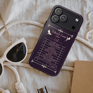 Op de afbeelding: Een donkerpaarse telefoonhoes met een bibliotheekkaartontwerp. De hoes heeft een lijst met titels en de tekst "Library Card" bovenaan. Een witte zonnebril en een crèmekleurige tas staan ook in de afbeelding.