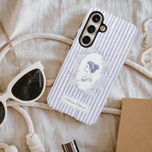 Puede incluir: Funda de teléfono con un patrón de rayas verticales en azul claro y blanco, con un retrato vintage de una mujer dentro de un marco floral. El texto "Allison Thorne" está debajo. Gafas de sol blancas y una cadena dorada también son visibles.
