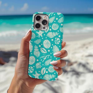 Op de afbeelding: Een turquoise telefoonhoesje met een herhaald patroon van witte schelpen, zeesterren en andere mariene elementen. De hoes wordt in een hand gehouden tegen een strandachtergrond met wit zand en blauwe oceaan.