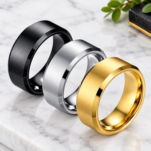 May include: Three metal rings arranged in a row, featuring a matte finish with polished beveled edges. The set includes one black ring, one silver-toned ring, and one gold-toned ring, each displaying a wide, flat band design.