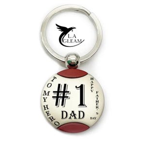 May include: Silver keychain with a round pendant. The pendant has a red border and features the text "#1 DAD" in black, with "TO MY HERO" and "HAPPY FATHER'S DAY" around the edge. The keychain has a silver ring and a small, round accent.