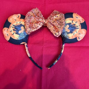 May include: Halloween-themed mouse ears headband. The black ears feature a spooky castle and bats design with orange and yellow accents. A sequined orange bow sits between the ears. The headband is black with a floral pattern.