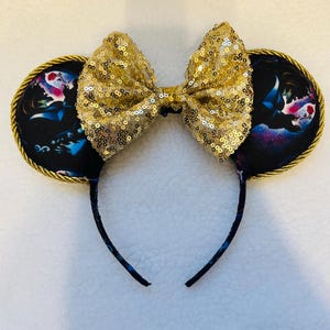 May include: Mouse ears with a gold sequin bow and black ears with a colourful design. The ears are trimmed with gold rope and attached to a black headband.
