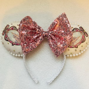 Puede incluir: Orejas de ratón con un gran lazo rosa con lentejuelas. Las orejas color crema tienen una imagen enmarcada de un castillo y están adornadas con perlas. La diadema es blanca con un diseño de encaje.