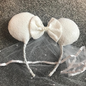 May include: A white sequined mouse ear headband with a matching bow. The headband includes a delicate veil decorated with small pearls, creating a whimsical and elegant accessory. Ideal for themed events or bridal wear.