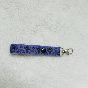 May include: A purple keychain strap with a black pattern of spooky mansion silhouettes. The strap is attached to a silver-colored metal clasp and a black snap. The background is a white, textured surface.