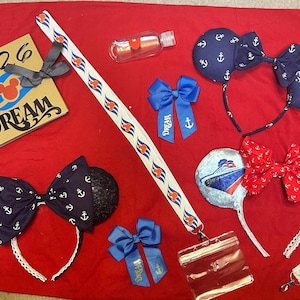 Puede incluir: Una colección de accesorios de temática náutica sobre un fondo rojo. Incluye diademas de orejas de ratón azul marino con patrones de anclas, un cordón, lazos, un cuaderno con el texto "2026 DREAM" y una botella pequeña.
