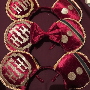 May include: Burgundy velvet mouse ear headbands with gold trim and accents. One pair features a bow, while others have gold glitter details and a gold emblem. The headbands are a festive accessory.