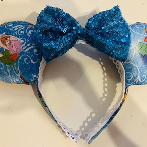 May include: Blue mouse ear headband with a sequined bow. The ears feature a light blue background with a cartoon fox and rabbit illustration. The headband has a white lace trim.