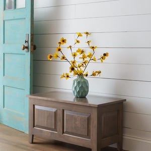 Peut inclure: Un coffre de rangement en bois brun moyen, avec deux panneaux encastrés sur le devant. Un vase turquoise contenant des fleurs jaunes est posé dessus. Le coffre est posé sur un sol en bois. Le coffre mesure environ 100 cm de large.