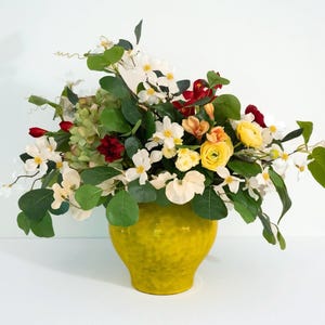 May include: A vibrant floral arrangement in a textured yellow vase. The bouquet features a mix of white, yellow, red, and orange flowers with green foliage. The vase is a bright, solid color, creating a striking contrast with the flowers.