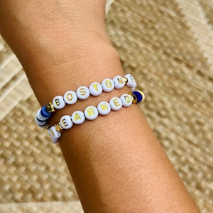 May include: Two beaded bracelets spelling "BOSTON" and "EARNED" in gold letters on white beads. The bracelets have gold star and blue accents. The bracelets are on a person's arm.