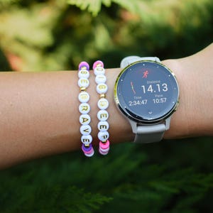 May include: Two beaded bracelets featuring white round beads with gold letters spelling BE BRAVE and DIG DEEP, accented with small gold spheres and pink, purple, and white flat circular beads, worn alongside a light gray digital sports watch.
