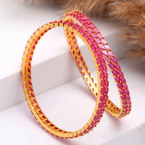 May include: Two gold bangles featuring a design of pink gemstones. The gemstones are closely set, forming a continuous band of colour. The bangles are presented on a white surface with a neutral backdrop.