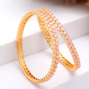 May include: Two gold-colored bangles with a delicate design. Each bangle features a row of small, light pink stones. The bangles are displayed on a white surface, showcasing their intricate details and elegant style.