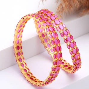 Könnte beinhalten: Zwei goldene Armreifen mit ovalen, rosa Edelsteinen. Die Armreifen sind gestapelt, was das detaillierte Design und die lebendige Farbe hervorhebt. Der Schmuck ist vor einem weißen Hintergrund platziert.