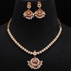 Può includere: Un set di collana e orecchini dorati. La collana presenta una fila di piccole perle bianche rotonde e un ciondolo con un design circolare, dettagli rossi e piccole perle bianche. Gli orecchini riprendono il design del ciondolo.