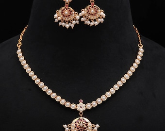 May include: A gold-toned necklace and earring set. The necklace features a strand of small, round, white pearls and a pendant with a circular design, red accents, and small white pearls. The earrings mirror the pendant design.