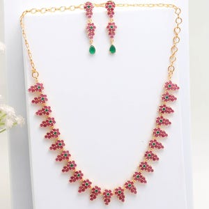 May include: A gold-toned necklace and earring set. The necklace and earrings feature clusters of small, red gemstones interspersed with green gemstones. The earrings have a teardrop-shaped green gemstone at the bottom.