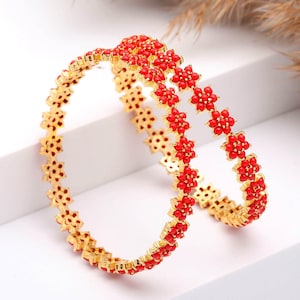 May include: Two gold bangles with a floral design. Each bangle is adorned with small, red flower-shaped accents. The bangles are displayed on a white surface, with a neutral background.