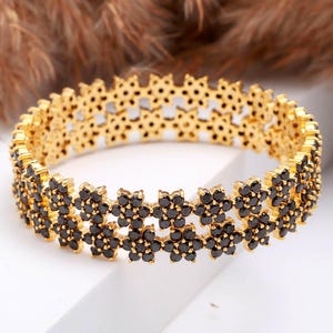 May include: Gold-toned bracelet featuring a floral design with black gemstones. The bracelet has a delicate, openwork pattern and is set against a neutral background. A stylish piece of jewelry.