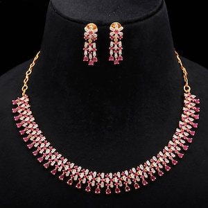 May include: A gold necklace and earring set on a black display. The necklace and earrings have a repeating pattern of red and white gemstones. The earrings are floral with three gemstone strands hanging down.