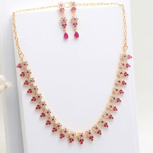 Puede incluir: Un conjunto de collar y pendientes dorados. El collar y los pendientes presentan un diseño floral con piedras preciosas rojas y transparentes. Los pendientes tienen una piedra preciosa roja en forma de lágrima en la parte inferior. Las joyas se exhiben sobre una superficie blanca.