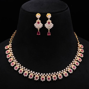 Könnte beinhalten: Ein goldfarbenes Halsketten- und Ohrring-Set. Die Halskette und die Ohrringe weisen ein florales Design mit roten und klaren Edelsteinen auf. Der Schmuck wird auf einem schwarzen Schaufensterkopf vor schwarzem Hintergrund präsentiert.