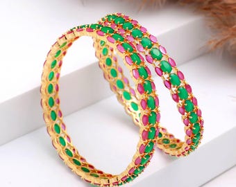Puede incluir: Dos brazaletes dorados con piedras preciosas ovaladas verdes y rosas alternas. Los brazaletes se exhiben sobre una superficie blanca, mostrando su intrincado diseño y colores vibrantes. Las piedras están engastadas juntas.