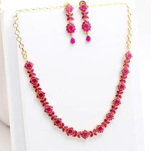 Peut inclure: Un ensemble collier et boucles d'oreilles de couleur dorée. Le collier et les boucles d'oreilles présentent un motif floral avec des pierres précieuses roses vibrantes. Les boucles d'oreilles sont de style pendantes, et le collier est de style ras du cou. Les bijoux sont exposés sur une surface blanche.
