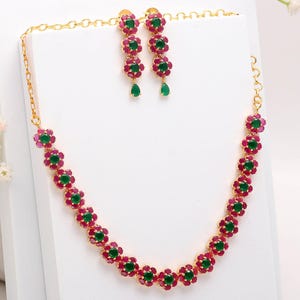 May include: A gold-coloured necklace and earring set. The necklace and earrings feature a floral design with alternating red and green gemstones. The earrings have a teardrop-shaped green gemstone at the bottom.