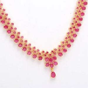 May include: A gold-coloured necklace adorned with pink gemstones. The oval-shaped stones are arranged in a floral design, with a teardrop pendant at the centre. The necklace is displayed against a white backdrop.