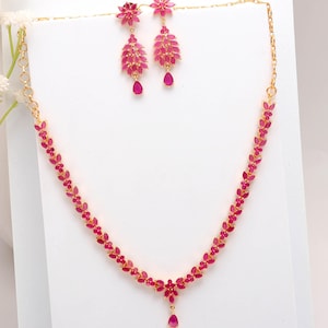 Puede incluir: Un conjunto de collar y pendientes dorados con piedras preciosas de color rubí. El collar tiene un diseño floral con un colgante en forma de lágrima. Los pendientes tienen una parte superior floral y un diseño en cascada con una gema en forma de lágrima.