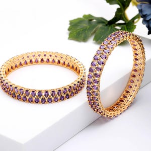 May include: Two gold bangles with purple gemstone accents. The bangles are set with oval-shaped purple gemstones. The gold bands have a textured design. The bangles are displayed on a white surface.