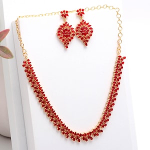 Puede incluir: Un conjunto de collar y pendientes dorados con piedras rojas. Los pendientes son colgantes y el collar es tipo gargantilla. El conjunto presenta un diseño floral y está expuesto sobre una superficie blanca.