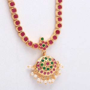May include: Gold-toned necklace featuring a series of red gemstones. The pendant has a circular design with red and green accents, and small white pearls. The necklace is a classic piece of jewelry.