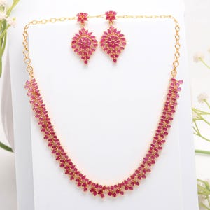May include: A gold-coloured jewellery set comprising a necklace and matching earrings. The necklace and earrings are embellished with many small, ruby-coloured gemstones. The earrings are teardrop-shaped, and the necklace curves elegantly around the neckline.