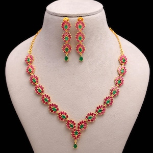 May include: A gold-toned necklace and earring set. The necklace and earrings feature a floral design with red and green gemstones. The earrings are dangling, and the necklace is displayed on a mannequin.
