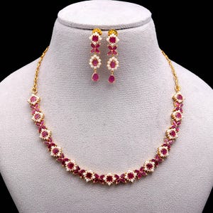 Puede incluir: Un collar dorado y un juego de pendientes a juego. El collar y los pendientes presentan un diseño floral con piedras de color rubí y pequeños detalles blancos. Los pendientes tienen un colgante de rubí en forma de lágrima.