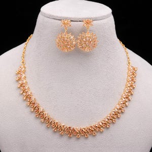 Puede incluir: Un conjunto de joyas de color dorado que incluye un collar y pendientes a juego. El collar y los pendientes están adornados con piedras en forma de lágrima de color melocotón y diseños redondos en forma de flor. El conjunto se exhibe sobre un busto de maniquí.